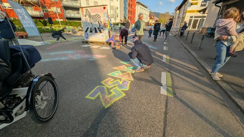 Eine Person kniet nieder und schreibt "ZEBRA", während andere in der Nähe laufen oder Rad fahren. Im Hintergrund sind Gebäude und ein helles Wandgemälde zu sehen.