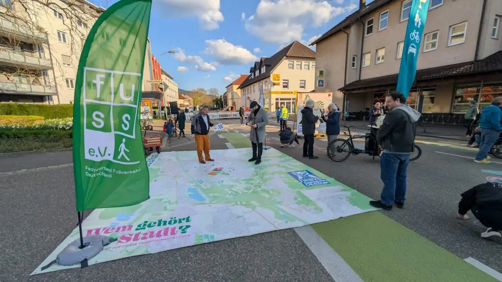 Menschen stehen und gehen um eine große Karte herum, die während einer Veranstaltung im Freien auf einer abgesperrten Straße ausgelegt ist. Im Hintergrund sind grüne Banner zu sehen, die für fußgängerfreundliche städtische Räume und Gebäude werben, während der Himmel blau und wolkenverhangen ist.
