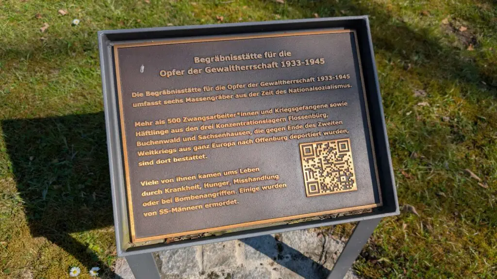 Eine Metalltafel in deutscher Sprache erklärt eine Begräbnisstätte für Opfer der nationalsozialistischen Gewalt (1933-1945), darunter Zwangsarbeiter und KZ-Häftlinge. Die Gedenktafel enthält auch einen QR-Code.