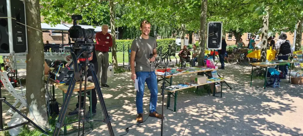 Ein Mann steht lächelnd mit einem Mikrofon vor einer Kameraeinstellung bei einer Veranstaltung im Freien unter Bäumen, mit Tischen, auf denen bunte Gegenstände und Menschen im Hintergrund zu sehen sind.