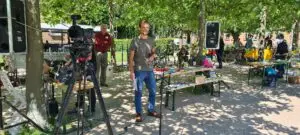 Ein Mann steht lächelnd mit einem Mikrofon vor einer Kameraeinstellung bei einer Veranstaltung im Freien unter Bäumen, mit Tischen, auf denen bunte Gegenstände und Menschen im Hintergrund zu sehen sind.