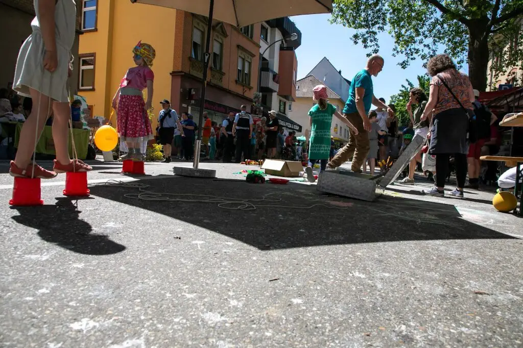 Kinder und Erwachsene vergnügen sich auf einer sonnigen Straße im Freien mit Spielen, Spielzeug und Kreidezeichnungen auf dem Boden. Einige Menschen stehen unter einem großen Regenschirm, während andere in der Nähe spazieren gehen oder zusehen. Im Hintergrund sind Gebäude und Bäume zu sehen.