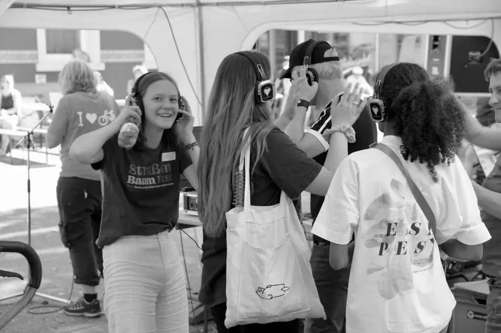 Eine Gruppe junger Leute mit Kopfhörern genießt eine Veranstaltung im Freien. Ein Mädchen lächelt und blickt in die Kamera, während andere sich gegenüberstehen und sich unterhalten. Die Szene ist lebhaft und zwanglos und findet unter einem Vordach statt.