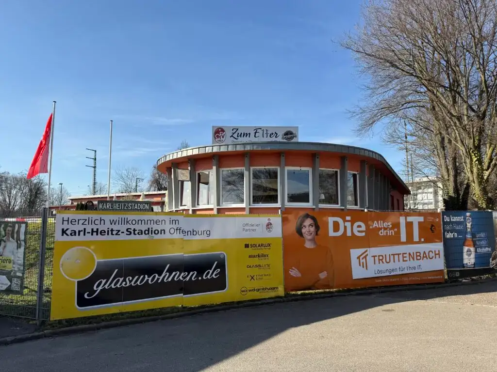 Ein rundes, orangefarbenes Gebäude mit der Aufschrift Zum Elfer steht hinter einem Zaun mit bunten Bannern, darunter Werbung und ein Willkommensschild für das Karl-Heitz-Stadion in Offenburg. In der Nähe stehen Bäume und eine rote Fahne.
