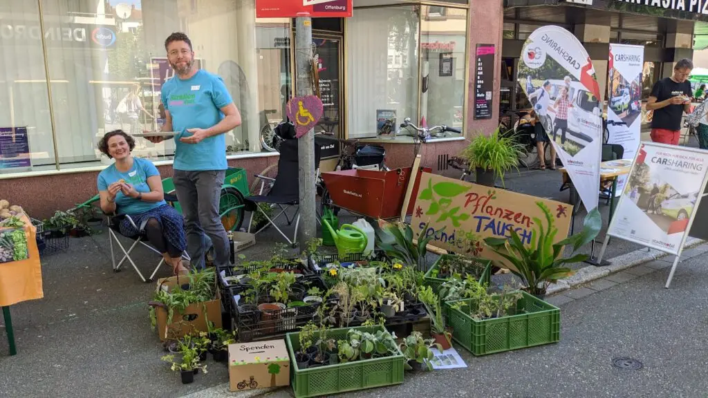 Zwei Personen sitzen und stehen hinter einem Pflanzentauschstand mit grünen Pflanzen in Kisten, einem bunten Schild mit der Aufschrift "Pflanzen Tausch" und Fahrrädern und Plakaten im Hintergrund auf einer sonnigen Straße.