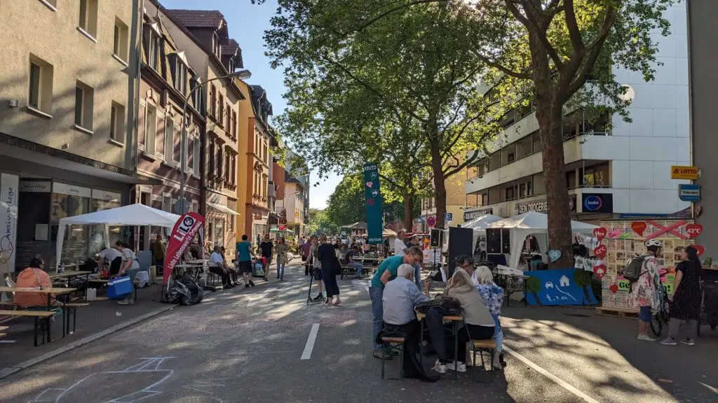 Ein lebhaftes Straßenfest mit Menschen, die an Tischen sitzen, spazieren gehen und Stände auf beiden Seiten einer geschlossenen, von Bäumen und Gebäuden gesäumten Stadtstraße an einem sonnigen Tag besuchen.