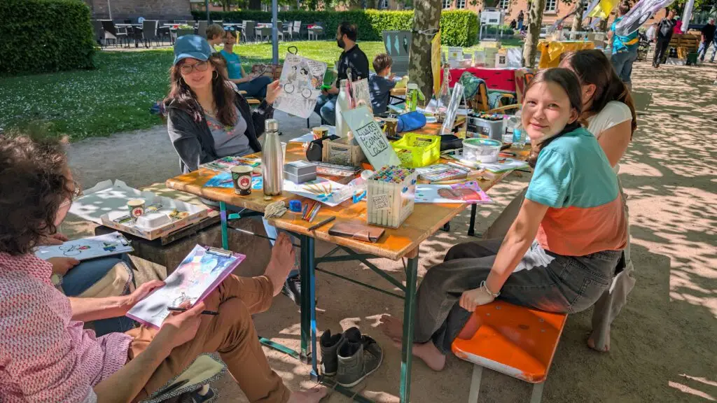 Menschen sitzen um einen Tisch im Freien und zeichnen und malen mit auf dem Tisch verstreutem Kunstzubehör. Einige lächeln und sind barfuß, genießen einen sonnigen Tag in einem Park, mit mehr Menschen und kreativen Aktivitäten im Hintergrund.