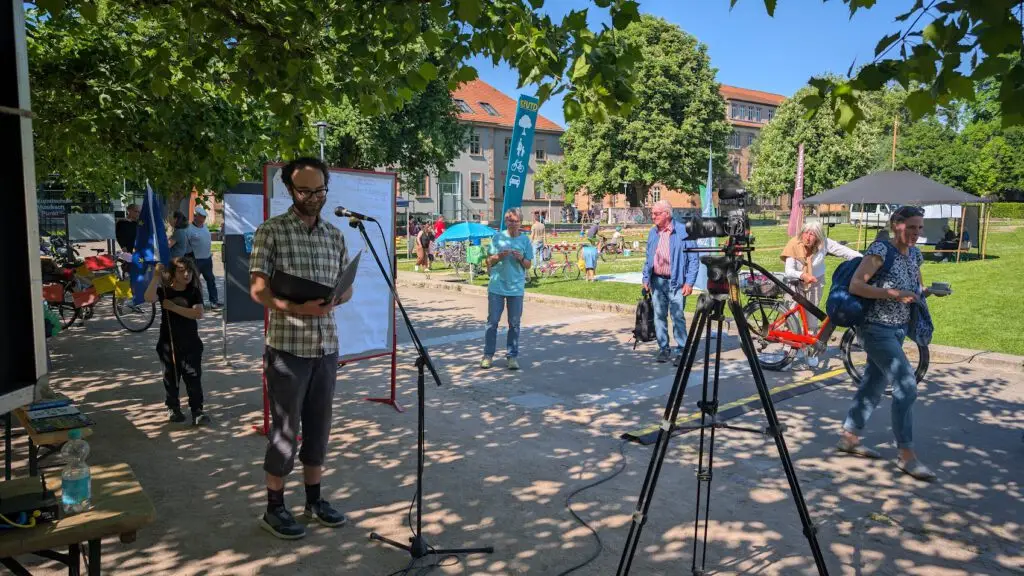 Ein Mann steht an einem Mikrofon und liest aus einer Mappe in einem sonnigen Park, in dessen Nähe Menschen spazieren gehen und sich unterhalten. Die Bäume werfen Schatten, und im Vordergrund ist eine Kamera aufgestellt. Im Hintergrund sind ein Spielplatz und Gebäude zu sehen.