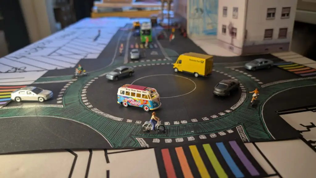 Ein Miniaturmodell eines Kreisverkehrs mit Spielzeugautos, einem gelben Lieferwagen, einem bunten Bus und kleinen Figuren von Menschen auf einer Straße mit einem regenbogenfarbigen Zebrastreifen und Gebäuden im Hintergrund.