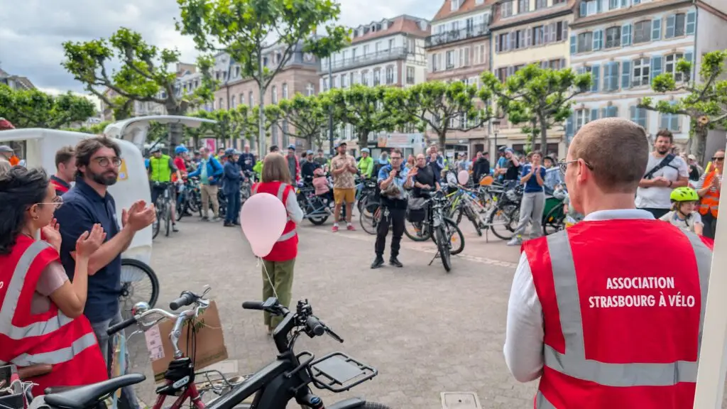 Auf einem europäischen Stadtplatz versammelt sich eine Menschenmenge im Freien, viele mit Fahrrädern. Die Menschen tragen rote Westen mit der Aufschrift Association Strasbourg à Vélo. Einige applaudieren, während eine Person mit einem Luftballon in der Mitte steht. Im Hintergrund sind Gebäude und Bäume zu sehen.