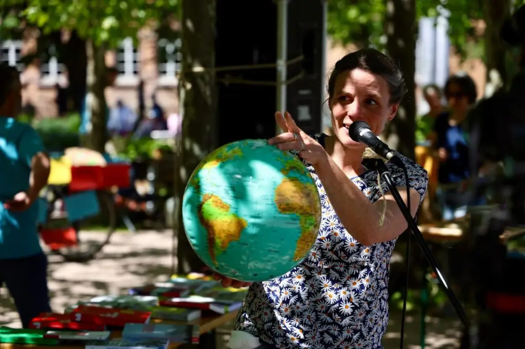 Eine Frau hält einen Globus und spricht in ein Mikrofon bei einer Veranstaltung im Freien, mit Tischen und Menschen im Hintergrund unter Bäumen.