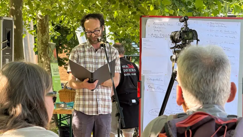 Ein Mann steht im Freien und liest aus einer Mappe in ein Mikrofon, während er sich dem Publikum zuwendet. Hinter ihm steht eine große Tafel mit Notizen und eine auf einem Stativ montierte Kamera. Die Bäume spenden Schatten und das Sonnenlicht fällt durch die Blätter.