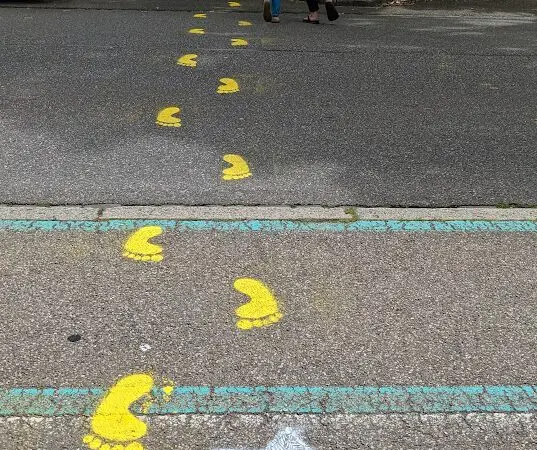 Gelb gemalte Tierspuren führen über eine Straße. Eine Zebra-Schablone und ein deutscher Text befinden sich auf der Straße. Im Hintergrund halten sich ein Erwachsener und ein Kind an den Händen, während sie den Spuren folgen. In der Nähe sind Bäume und Autos zu sehen.