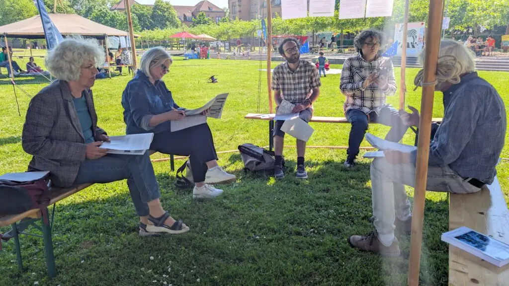 Fünf Erwachsene sitzen im Kreis auf Bänken und Stühlen im Gras, halten Papiere in der Hand und unterhalten sich. Es ist ein sonniger Tag, und im Hintergrund sind Bäume, Zelte und Menschen zu sehen, was auf einen Park oder eine Gemeinschaftsveranstaltung hindeutet.