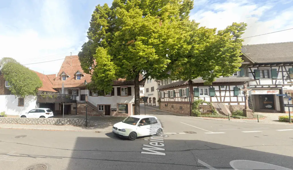 Ein kleines weißes Auto fährt an einem sonnigen Tag durch eine Kreuzung, die von traditionellen Fachwerkhäusern und grünen Bäumen gesäumt ist. Auf dem Straßenschild steht "Weingartenstraße".