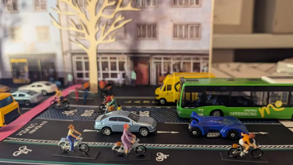 Eine Miniaturstadtszene mit Modellautos, Bussen und Radfahrern auf einer Straße. Im Hintergrund befinden sich kleine Gebäude, ein weißer Baum und verschiedene Fahrzeuge, darunter ein grüner Bus und ein gelber Lieferwagen.