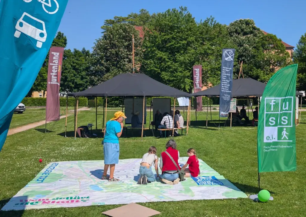 An einem sonnigen Tag versammeln sich Menschen im Freien, einige sitzen an Tischen unter Zelten, andere stehen oder knien auf einer großen Karte, die auf dem Rasen ausgelegt ist. In der Nähe sind bunte Transparente angebracht, und im Hintergrund stehen Bäume.