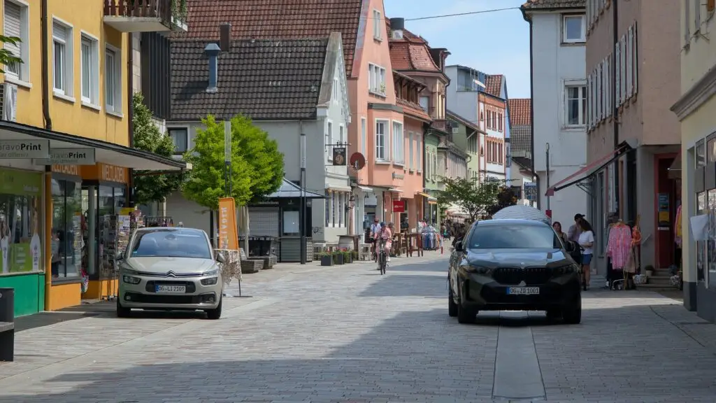 Eine ruhige europäische Straße mit farbenfrohen Gebäuden, auf beiden Seiten geparkten Autos, Spaziergängern und Radfahrern und Geschäften, die die Kopfsteinpflasterstraße unter einem klaren Himmel säumen.