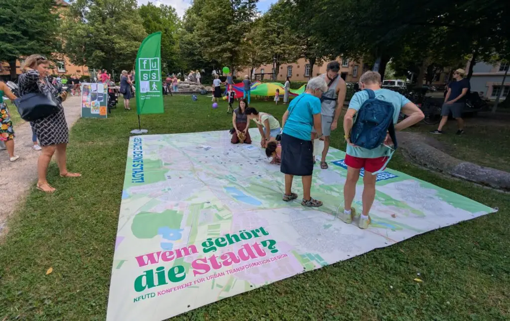 Menschen betrachten eine große Karte, die bei einer Veranstaltung im Freien in einem Park auf dem Rasen ausgelegt ist. Auf der Karte steht "Wem gehört die Stadt?", im Hintergrund sind Informationsbanner und andere Teilnehmer zu sehen.