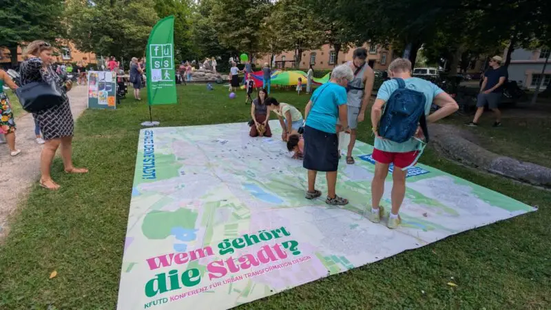 Menschen betrachten eine große Karte, die bei einer Veranstaltung im Freien in einem Park auf dem Rasen ausgelegt ist. Auf der Karte steht "Wem gehört die Stadt?", im Hintergrund sind Informationsbanner und andere Teilnehmer zu sehen.