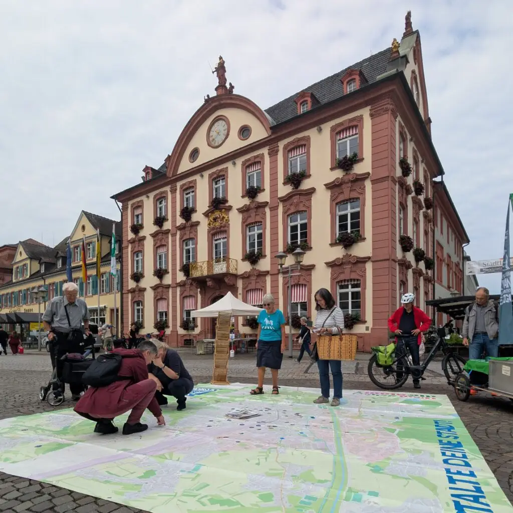 Mehrere Personen beschäftigen sich mit einer großen Landkarte, die auf dem Kopfsteinpflaster eines Stadtplatzes vor einem verschnörkelten, historischen Gebäude mit roten Zierleisten und Blumenkästen unter den Fenstern liegt. Einige Leute sind auf Fahrrädern unterwegs.