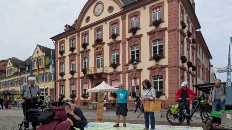 Mehrere Personen beschäftigen sich mit einer großen Landkarte, die auf dem Kopfsteinpflaster eines Stadtplatzes vor einem verschnörkelten, historischen Gebäude mit roten Zierleisten und Blumenkästen unter den Fenstern liegt. Einige Leute sind auf Fahrrädern unterwegs.