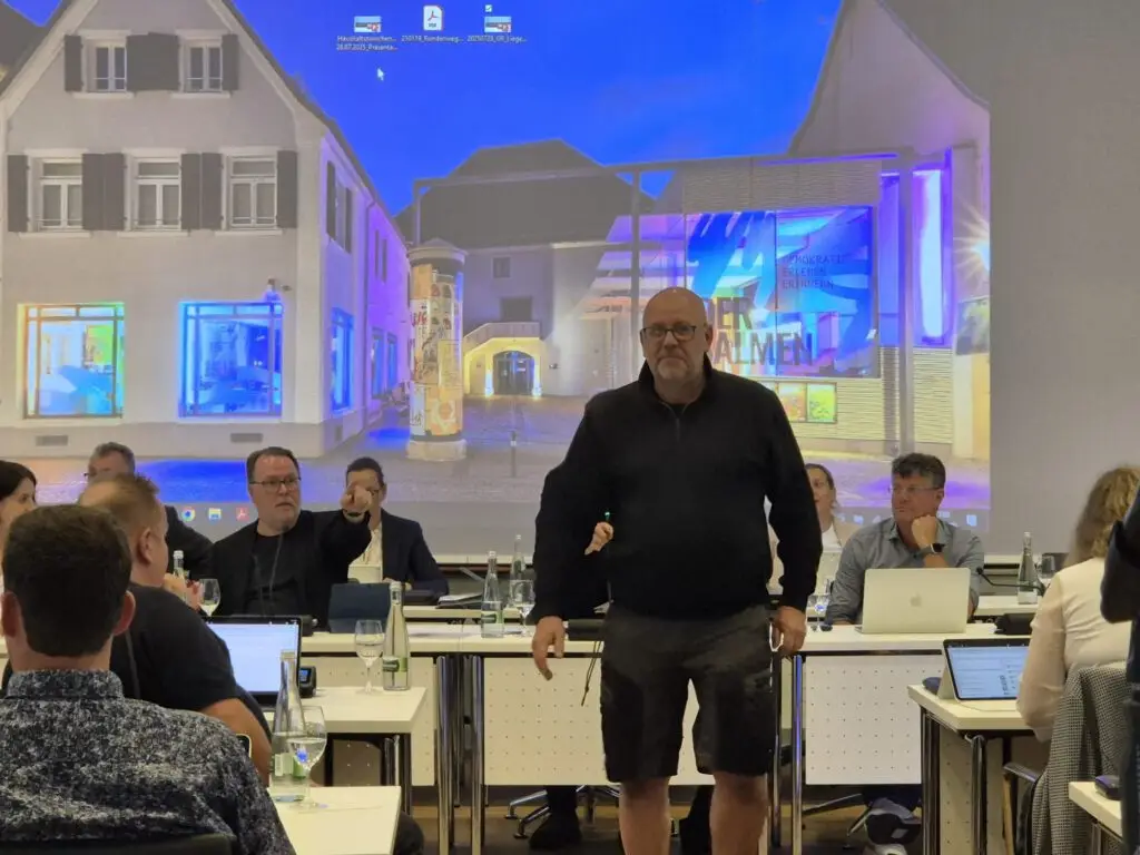 Ein Mann in schwarzem Hemd und kurzen Hosen steht vor einer Gruppe von Menschen, die an Tischen mit Laptops und Flaschen sitzen. An der Wand hinter ihnen ist eine bunte Gebäudeszene projiziert.