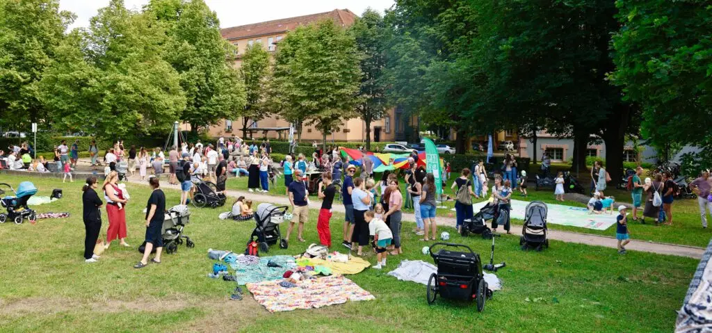 Eine große Gruppe von Menschen, darunter Familien mit Kindern und Kinderwagen, versammelt sich in einem grünen Park. Einige Kinder spielen mit einem bunten Fallschirm, während andere sich auf Decken ausruhen. Hohe Bäume und Gebäude umgeben den Park an einem sonnigen Tag.