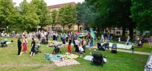 Eine große Gruppe von Menschen, darunter Familien mit Kindern und Kinderwagen, versammelt sich in einem grünen Park. Einige Kinder spielen mit einem bunten Fallschirm, während andere sich auf Decken ausruhen. Hohe Bäume und Gebäude umgeben den Park an einem sonnigen Tag.