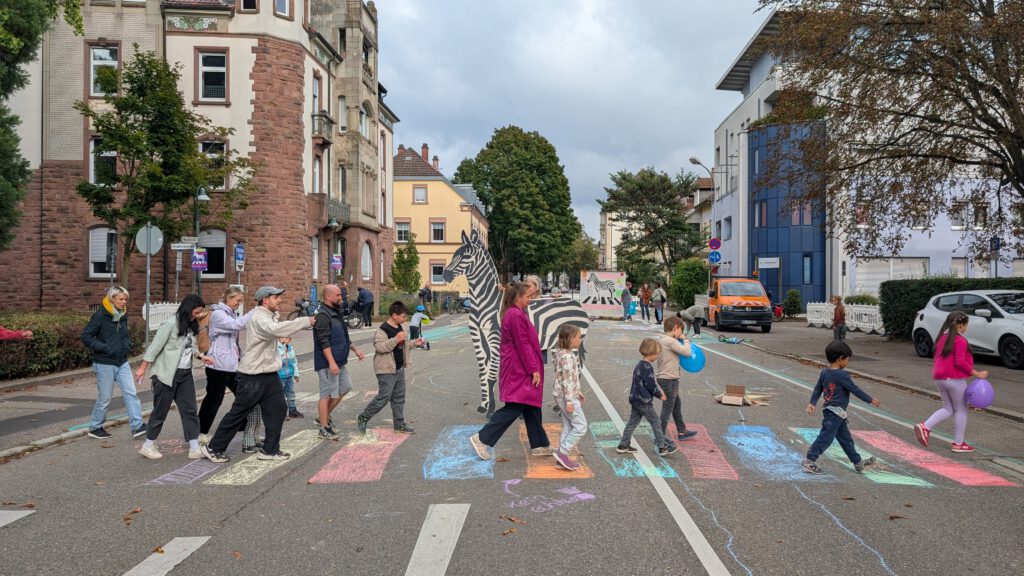 Eine Gruppe von Menschen, darunter auch Kinder, überquert eine mit bunten Kreidestreifen bemalte Straße, die einem Zebrastreifen ähnelt. Gebäude, Bäume und geparkte Autos säumen den Hintergrund, und eine Person in einem Zebrakostüm steht in der Nähe der Gruppe.