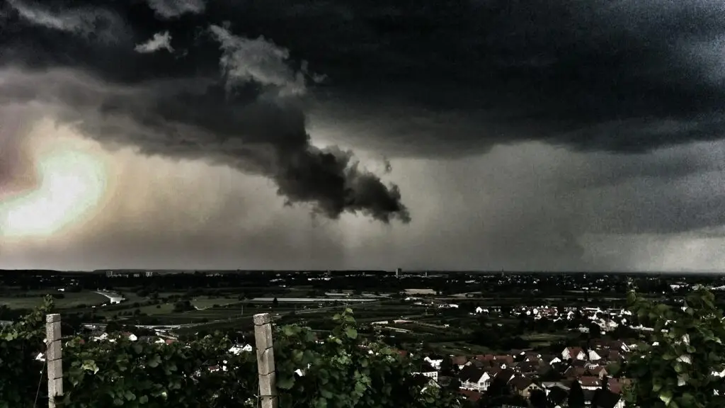 Dunkle Gewitterwolken ziehen über einer Stadt und den Feldern auf. In der Ferne fällt starker Regen, und ein dunkler, dramatischer Himmel beherrscht die Szene.