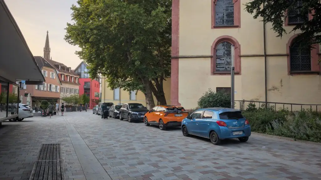 Autos parken auf einer Kopfsteinpflasterstraße neben einem großen gelben Gebäude mit gewölbten Fenstern; Bäume spenden Schatten, und in der Ferne gehen Menschen an weiteren Gebäuden vorbei. Im Hintergrund ist eine Kirchturmspitze zu sehen.