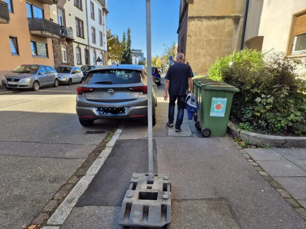 Ein Mann geht an einem sonnigen Tag in einem Wohngebiet auf einem Gehweg, der teilweise durch einen Pfosten und einen großen Sockel versperrt ist, mit einer grünen Mülltonne und geparkten Autos in der Nähe.