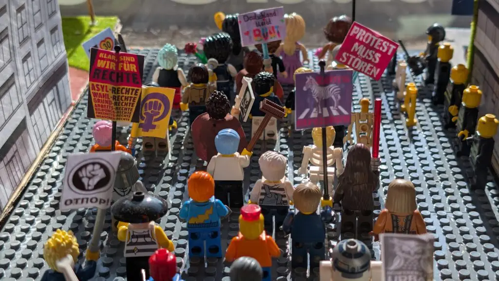 Eine Gruppe von LEGO Minifiguren hält Protestschilder in deutscher Sprache und Symbole für die Rechte der Frauen in der Hand, während sie auf einer von LEGO nachgebauten Straße marschieren und so eine lebhafte Demonstrationsszene simulieren.