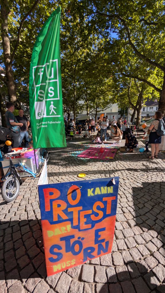 Auf einem farbenfrohen Protestschild auf einem kopfsteingepflasterten Platz steht "Protest kann störend sein, muss aber sein". In der Nähe versammeln sich Menschen unter grünen Bäumen, mit einer großen grünen Flagge im Hintergrund.