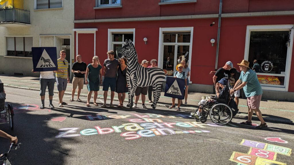 Eine Gruppe von Menschen, darunter eine Person im Rollstuhl, steht auf der Straße und hält Zebrastreifen und einen Zebrastreifen in der Hand. Vor ihnen sind bunte Kreidebilder und Himmel und Hölle auf den Bürgersteig gemalt.
