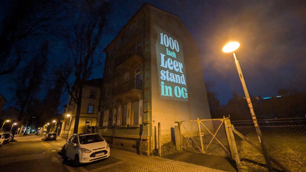 Eine nächtliche Straßenszene zeigt ein beleuchtetes Gebäude, auf dessen Seite die Aufschrift 1000 fach Leerstand in OG projiziert ist. Die Straßenlaternen in der Nähe beleuchten geparkte Autos und eine Toreinfahrt.