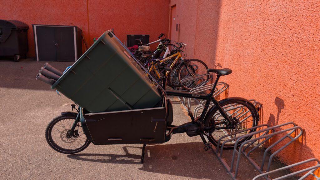 Ein Lastenfahrrad mit einer großen grünen Tonne im vorderen Fach ist neben mehreren anderen Fahrrädern an einer orangefarbenen Wand geparkt, in deren Nähe sich leere Fahrradständer befinden.