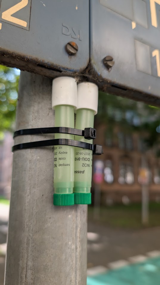 Zwei Probenröhrchen mit grünem Deckel sind mit schwarzen Kabelbindern an einer Metallstange befestigt. Die Röhrchen sind beschriftet und stehen senkrecht. Im Hintergrund sind ein unscharfes Gebäude und Bäume zu sehen.