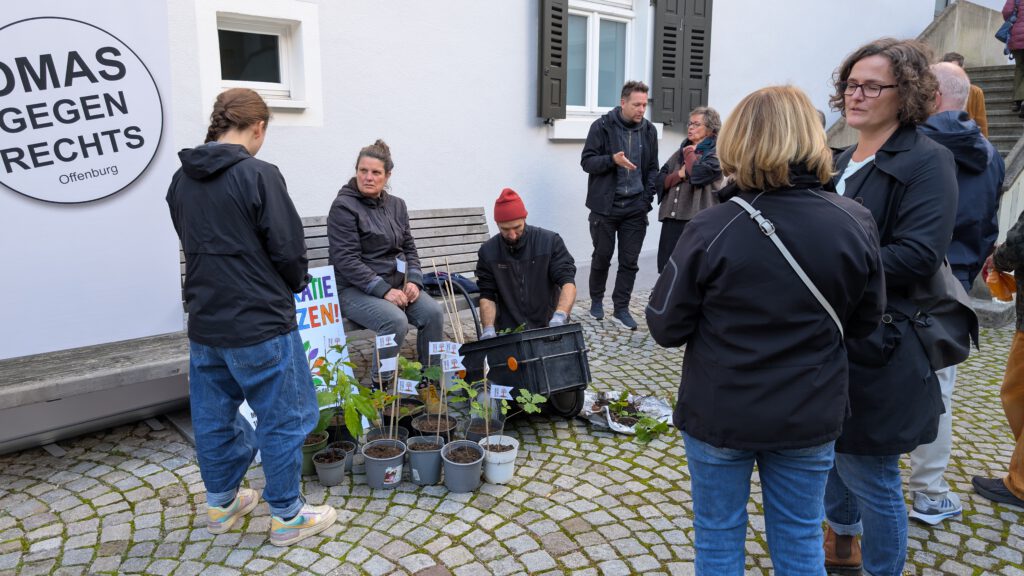 Eine Gruppe von Menschen versammelt sich im Freien auf einer gepflasterten Fläche in der Nähe eines Gebäudes. Topfpflanzen und kleine Schilder stehen auf dem Boden. Auf einem großen Schild an der Wand steht TOMAS GEGEN RECHTS. Einige Leute unterhalten sich, während andere die Pflanzen beobachten.