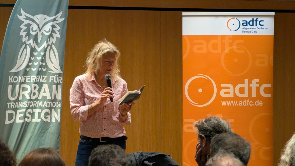 Eine Frau mit langen blonden Haaren spricht in ein Mikrofon, während sie auf der Bühne zwischen Bannern für eine Konferenz zum Thema Stadtumbau und den ADFC vor einem Publikum aus einem Buch liest.