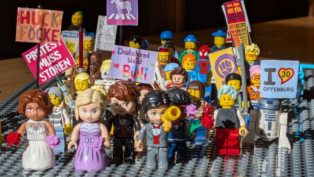 Eine Gruppe von LEGO Minifiguren, einige in formeller Kleidung, hält bunte Protestschilder in deutscher Sprache, darunter Regenbogenfahnen und Herzen. R2-D2 und andere Figuren in Kostümen stehen zwischen ihnen auf einer LEGO Grundplatte.