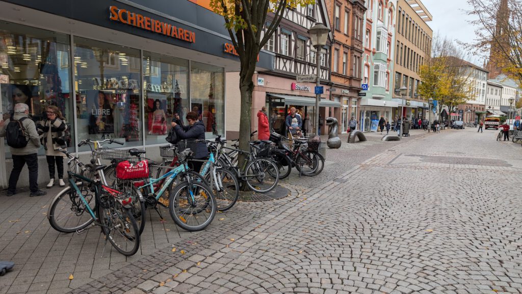 Mehrere Fahrräder sind entlang einer Fußgängerzone mit Kopfsteinpflaster in der Nähe eines Geschäfts mit der Aufschrift "SCHREIBWAREN" abgestellt. Die Menschen gehen spazieren, kaufen ein und unterhalten sich an einem bewölkten Tag unter herbstlichen Bäumen neben den Schaufenstern.