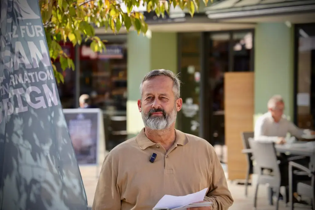 Uli Albicker. Ein Mann mit grauem Haar und Bart steht im Freien, hält Papiere in der Hand und trägt ein beigefarbenes Hemd mit einem angeklippten Mikrofon. Hinter ihm sitzen verschwommene Menschen an Tischen, und über ihm hängen Blätter von einem Baum.
