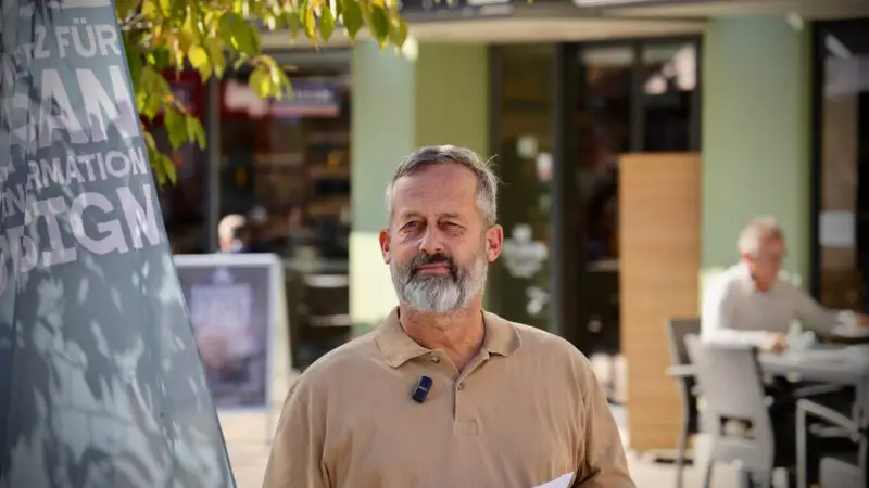 Ein Mann mit grauem Haar und Bart steht im Freien, hält Papiere in der Hand und trägt ein beigefarbenes Hemd mit einem angeklippten Mikrofon. Hinter ihm sitzen verschwommene Menschen an Tischen, und über ihm hängen Blätter von einem Baum.