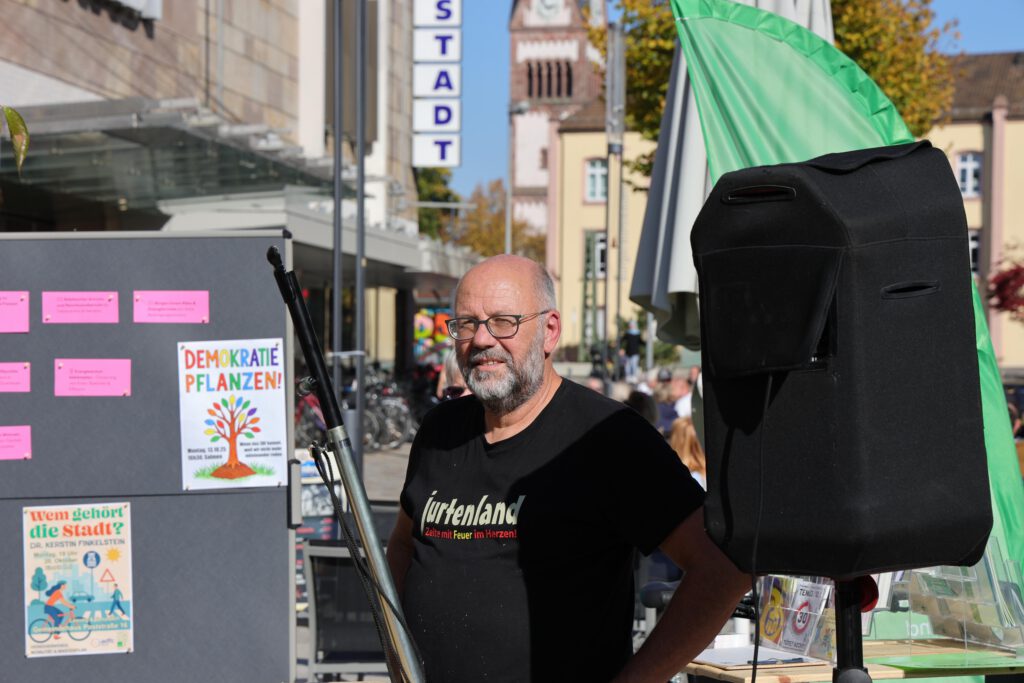 Ralph Fröhlich. Ein Mann mit Brille und Bart steht neben einem großen Lautsprecher und Informationsplakaten bei einer Veranstaltung im Freien. An einem sonnigen Tag sind im Hintergrund Gebäude und ein Uhrenturm zu sehen.