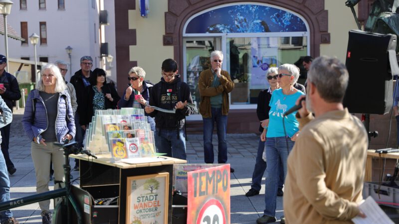 Eine Gruppe von Menschen steht und hört einem Mann zu, der bei einer Veranstaltung im Freien in ein Mikrofon spricht. In der Nähe liegen auf einem Tisch Bücher und Broschüren aus, und auf einem Schild ist ein Symbol für Tempo 30 zu sehen.