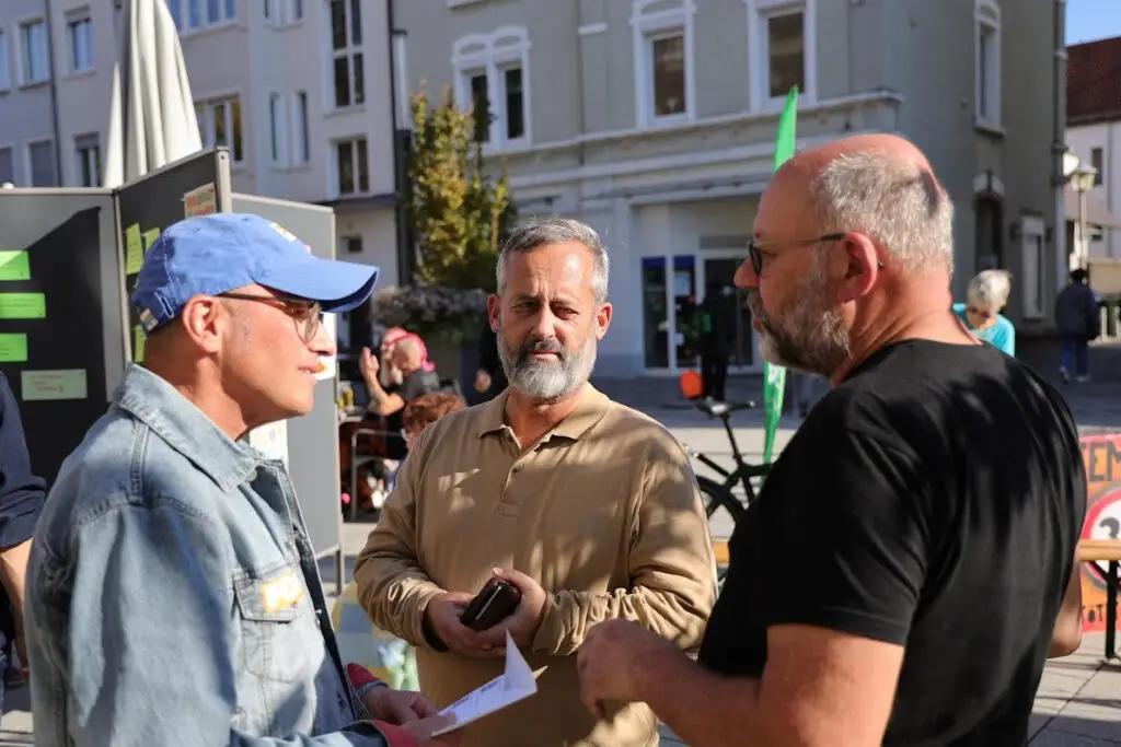 Armin Schmeling, Uli Albicker und Ralph Fröhlich tauschen sich über Bürgerbeteiligung aus. Drei Männer stehen an einem sonnigen Tag im Freien und unterhalten sich; einer trägt eine blaue Kappe und eine Sonnenbrille, ein anderer hält ein Telefon in der Hand, und der dritte hat eine Glatze und eine Brille. Im Hintergrund sind Gebäude und Menschen zu sehen.