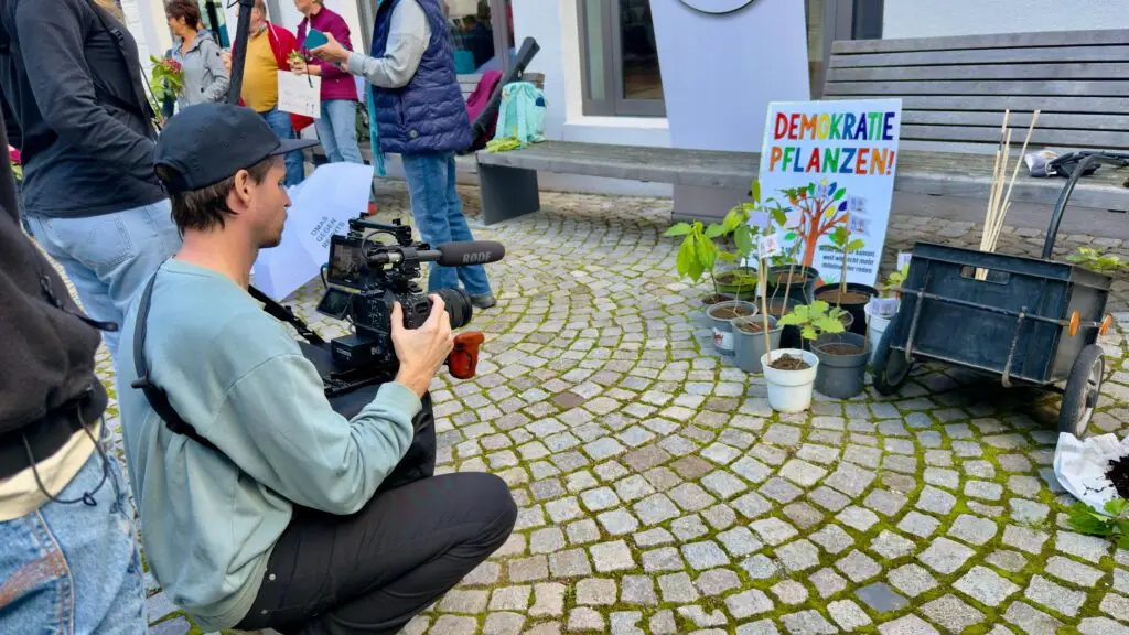 Ein Mann filmt Topfpflanzen und ein buntes Schild mit der Aufschrift Demokratie Pflanzen! bei einer Veranstaltung im Freien auf einer Kopfsteinpflasterstraße, im Hintergrund sind andere Menschen und eine Bank zu sehen.