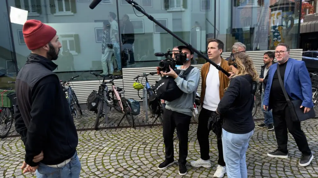 Ein Mann mit roter Mütze wird von einem Kamerateam mit einem Galgenmikrofon auf einer Kopfsteinpflasterstraße gefilmt, während drei Personen in der Nähe stehen, hinter ihnen sind mehrere Fahrräder geparkt.